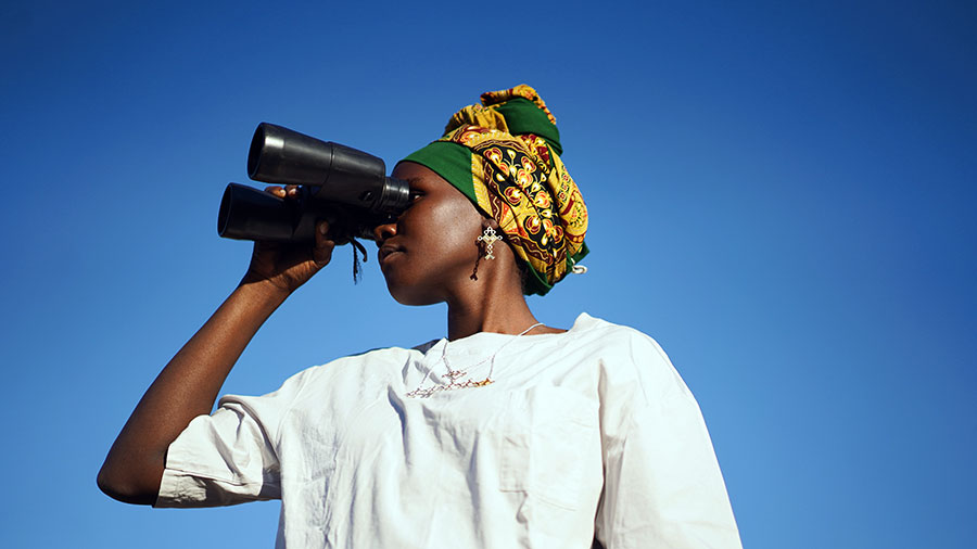 Woman in Tanzania in white long sleeve shirt and green, yellow headscarf looking through binoculars