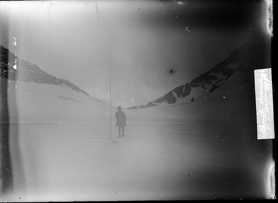 Black and white blurry photo of Axel Hamberg with a snow accumulation measurement instrument, standing between snowy hills, August 1899, Lullihavagge, Sarek, Sweden