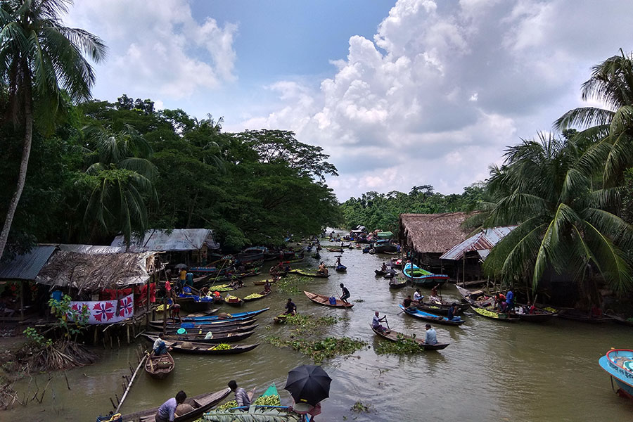 People move guavas across a river between wooden buildings on canoes. Someone with an umbrella sits on a boat at the centre of the image