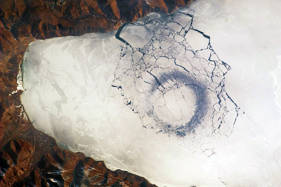 An areal photo of a dark circle amongst the cracking ice of a massive lake