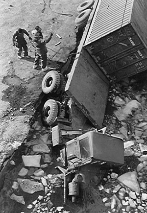 A black and white image shows three dock workers on the edge of an ice shelf. In front of them, a truck towing crates of cargo sinks into water surrounded by shards of ice