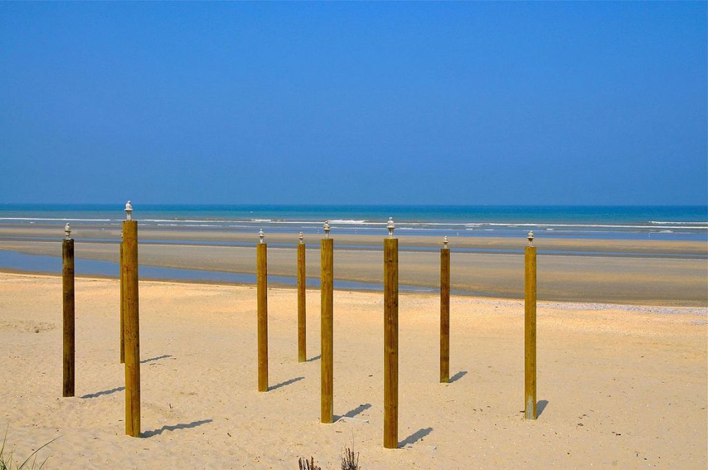 Photo of small figures standing on stilts on a sunny beach looking out towards the sea and blue sky